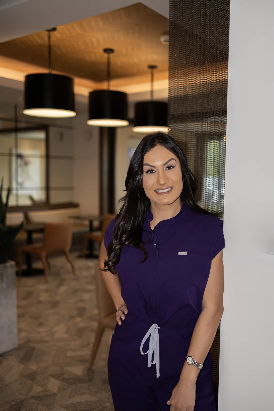 Bianca in plum scrubs, leaning on a column in a warm lobby