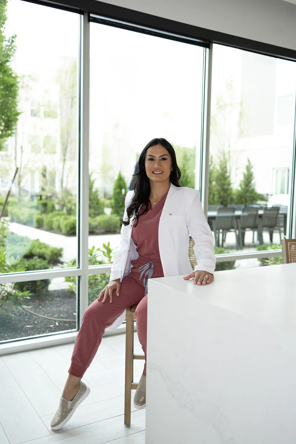 Bianca in a white coat, seated on a stool by tall windows